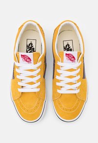 Žluté semišové tenisky s bílými tkaničkami, které mají fialový boční pruh a červenou značkovou nálepku Vans na jazyku. Gumová podrážka s detaily prošívání.