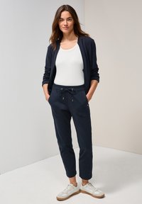 Marineblaue Zip-Jacke, weißes Tanktop und marineblaue Jogginghosen mit Kordelzug. Die Schuhe sind hellfarbene Sneakers mit braunen Sohlen.