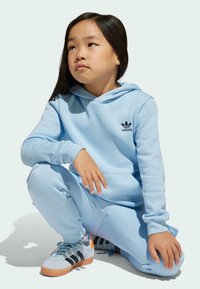 Svijetloplava kapuljača i trenirke od mekog materijala, s izvezenim crnim Adidas logotipom, u kombinaciji sa sivim i crnim tenisicama.