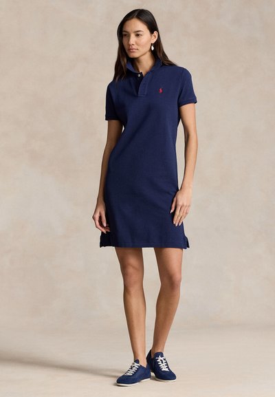 Granatowa sukienka polo z krótkimi rękawami, kołnierzykiem na dwa guziki, bocznymi rozcięciami oraz czerwonym logo na piersi. Noszona z niebieskimi trampkami.