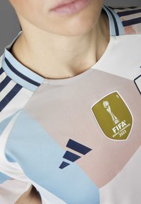 Camiseta de Adidas con un diseño geométrico en colores pastel, con cuello redondo, tres franjas en azul marino y un escudo de Campeones Mundiales FIFA 2023.