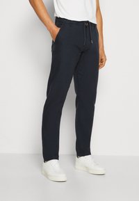 Dark navy trousers made of lightweight fabric. Features a drawstring waist, side pockets, and a slim fit. Paired with white sneakers.