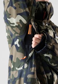Camouflagejack met een mix van groene, bruine en beige patronen. Opvallende oranje rits en een gaasvoering zichtbaar door de open zak.