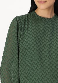 Haut vert texturé présentant un motif géométrique en chevron, avec un col rond et des manches bouffantes transparentes. Tissu doux avec une apparence superposée.