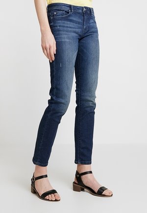 Persoon die een slimfit blauwe jeans en zwarte sandalen met enkelriempje en blokhak draagt, staand tegen een effen lichte achtergrond.