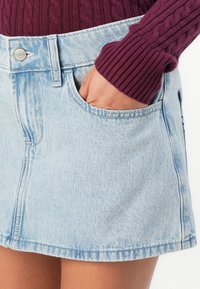 Hånd i lommen på en lyseblå denimskjørt, sammen med en vinrød kabelstrikket genser hvor ermet er synlig på håndleddet.