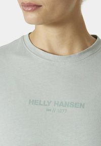 Světle šedé tričko s kulatým výstřihem a krátkými rukávy. Tištěný text v tmavší šedé barvě: "HELLY HANSEN HH // 1877" na přední straně.