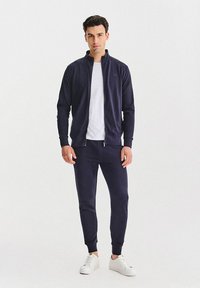 Granatowy dres z bluzą zapinaną na zamek i zwężanymi spodniami, wykonany z miękkiej tkaniny. Całość uzupełnia biała koszulka i białe trampki.