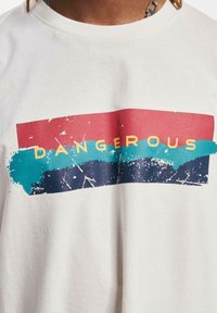 Krátký rukáv smetanového trička se multikolorovou grafikou se červenými, tyrkysovými a námořnickými pruhy a nápisem "DANGEROUS" v tučných žlutých písmenech.