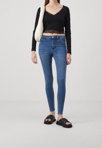 Top negro de manga larga recortado con borde de encaje, vaqueros ajustados de cintura alta azules y sandalias negras sin tacón. Diseño simple y ajustado.