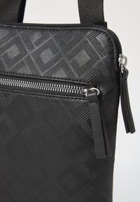 Black textured bag with geometric pattern, zip closure, and a front pocket. Features smooth and embossed materials with silver hardware accents.