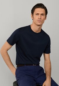Jeune homme portant un t-shirt bleu marine foncé et un pantalon bleu à fines rayures, assis avec une main dans sa poche sur un fond gris uni.
