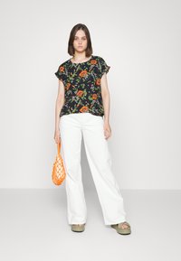 T-shirt floral preta com mangas curtas, combinada com calças largas brancas e sandálias verdes, segurando uma bolsa de rede laranja.