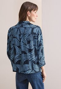 Chemise à manches longues à motifs bleus et noirs, au fit ample, avec des lignes courbes et un col. Associée à un jean en denim foncé.