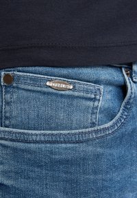 Zbliżenie na niebieską kieszeń dżinsów z denimu z srebrną odznaką marki Petrolino i fragmentem czarnej koszuli nad paskiem.