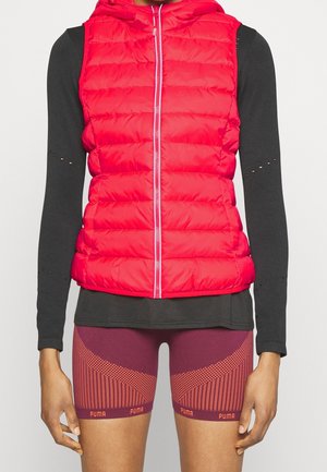 Persona delgada que lleva una chaqueta acolchada sin mangas de color rojo brillante sobre una camiseta negra de manga larga y pantalones cortos Puma color granate con detalles estampados.