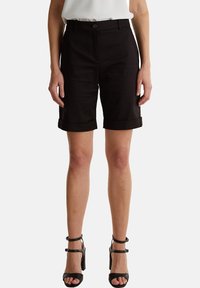 Shorts hitam yang dipotong dengan hem dilipat, terbuat dari kain halus, dilengkapi dengan penutupan tombol di depan dan loop sabuk. Dipadukan dengan sandal hak tinggi hitam.