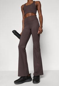 Smeđi atletski crop top i visoki flared tajice, izrađeni od rastezljivog materijala, u kombinaciji s crnim tenisicama i crnom bocom za vodu.