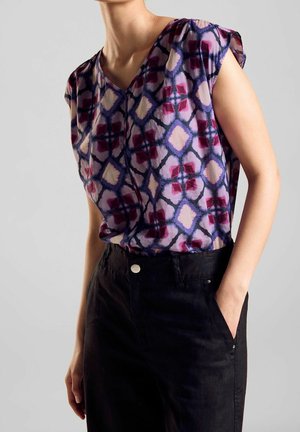 Kobieta w fioletowej, bezrękawnikowej bluzce w wzory, wpuszczonej w czarne spodnie z wysokim stanem, z jedną ręką w kieszeni, na jasnym, gładkim tle.