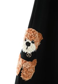 Prenda negra con un aplique de peluche de nutria texturizado en marrón y crema en la manga, con detalles faciales bordados y un logo en su camiseta.