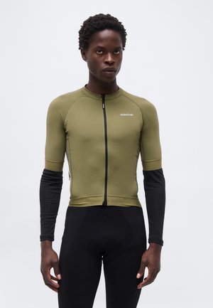 Άτομο που φοράει πράσινη ελιά κοντομάνικη μπλούζα ποδηλασίας με μαύρα μακριά μανίκια από μέσα και μαύρο σορτς ποδηλασίας, στέκεται μπροστά σε λευκό φόντο.