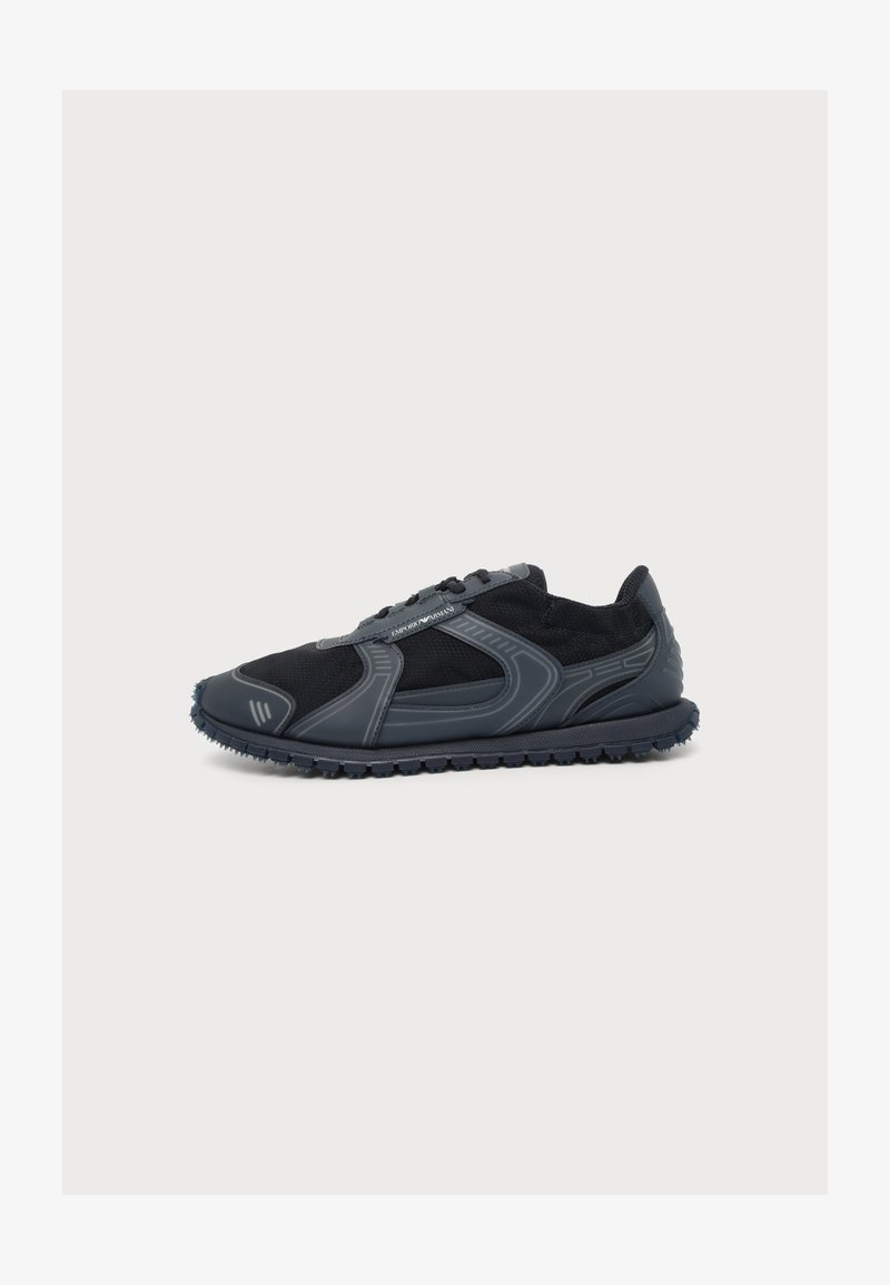 Μαύρο και σκούρο γκρι χαμηλό sneaker με υφή στην σόλα και πάνελ από δίχτυ, ετικέτα λογότυπου στο πλάι και στρογγυλεμένη μύτη σε λευκό φόντο.
