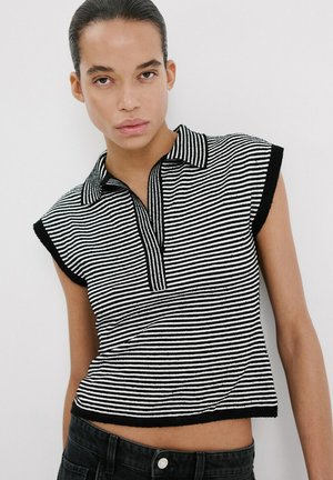 Joven con el cabello peinado hacia atrás, que lleva una camiseta sin mangas con cuello a rayas blancas y negras y pantalones negros, contra un fondo blanco.