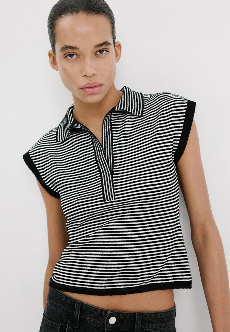 Ung person med bagstrøget hår iført en sort og hvidstribet ærmeløs skjorte med krave og sorte bukser mod en hvid baggrund.