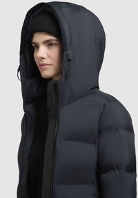 Tmavomodrá nadýchaná bunda s vysokým golierom, nastaviteľnou kapucňou a čiernym zipsom vpredu. Má prešívanú textúru a výrazné logo na strane.