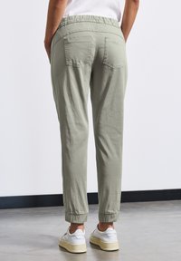 Ljust gröna tapered byxor med elastisk midja, uppvikta fållar och två bakfickor, i kombination med vita sneakers på ett betonggolv.