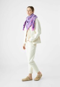 Witte denimjack, lichtbeige top, witte broek en tan klompen. Paarse sjaal met een geometrisch patroon om de nek gedrapeerd. Eenvoudige achtergrond.