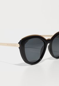 Occhiali da sole oversize neri con lenti grigie, dettagli dorati sulla montatura e una finitura lucida e brillante. Nome del marchio inciso sul braccio.