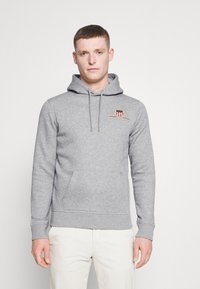 Grå hoodie med kengurulomme, snøre i hætten og broderet emblem. Blødt stof med ribbede manchetter og kant.