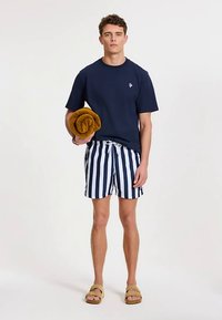 Granatowy bawełniany T-shirt, pasiasty strój kąpielowy w granacie i bieli, beżowe sandały, trzymający zwinięty żółty ręcznik. Prosty design, casualowy krój.