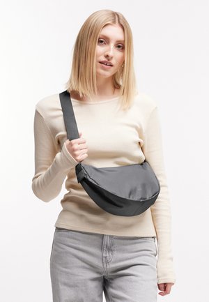 Jonge vrouw die een beige trui en grijze spijkerbroek draagt, met een grijze crossbody-tasriem over haar borst tegen een witte achtergrond.