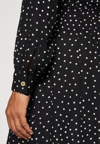 Zwart polkadotjurk met een knoopmanchet, lange mouwen en een licht doorschijnende stof, een getailleerde corsage en een vloeiende rok.