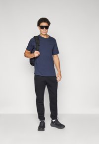 Marineblå kortermet T-skjorte, svarte joggebukser og svarte sneakers. Modellen bærer en svart ryggsekk og har på seg mørke solbriller.
