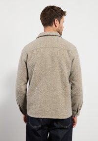 Veste d'extérieur texturée beige, floue, avec un col et des poignets boutonnés. Le design présente une coupe décontractée et un ourlet légèrement arrondi.