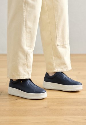 Personne portant un pantalon beige et des baskets slip-on bleu marine avec semelles blanches, debout sur un sol en bois.
