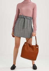 Lyserød rullekravetrøje, grå og sort ternet nederdel med bælte i taljen, sorte slip-on sko og en stor brun lædertaske.