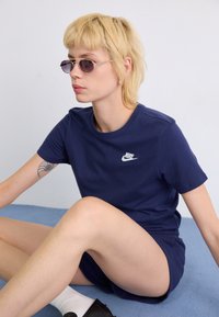 T-shirt en coton bleu marine avec un logo Nike blanc. Le modèle porte un short bleu marine et des lunettes de soleil rondes, assis sur une surface bleue.