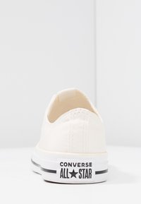 Baskets en toile de couleur crème avec un design bas, un talon arrondi, une semelle en caoutchouc et un logo noir « Converse All Star » sur le talon.