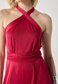 Rød halterneck-kjole med samlet bærestykke og talje. Glatt stoff, draperingsdesign og myk tekstur. Krydset stropper ved halsen.