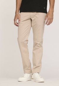 Pantalon en coton beige avec une coupe slim, dotés de poches avant et d'une texture lisse, porté avec des baskets blanches.