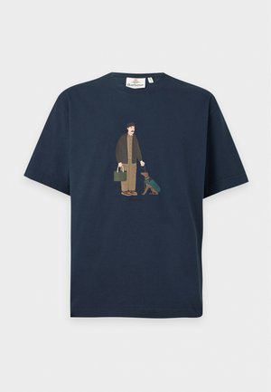 Un mannequin masculin porte un T-shirt bleu marine à motif graphique, une casquette olive, un jean foncé, une ceinture marron et des chaussures, sur un fond blanc uni.