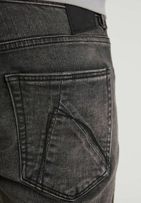 Jeans in denim grigio scuro con una finitura texture, caratterizzati da una tasca posteriore distintiva e dettagli di cucitura sottili, completati da una patch in pelle nera sulla vita.