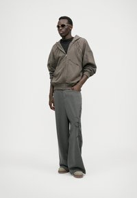 Grå lynlåse hættejakke, sort t-shirt, stribet vide bukser med broderi og grå fodtøj. Accessories inkluderer lys solbriller.