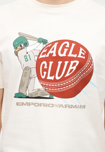 Κουτσομπολιά παπιού σε εξοπλισμό κρίκετ που χτυπάει με ρακέτα μια μεγάλη κόκκινη μπάλα με την επιγραφή «Eagle Club» και το κείμενο «Emporio Armani» από κάτω.