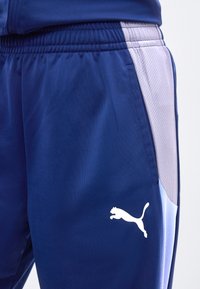 Κοντινό πλάνο σε μπλε και ανοιχτό γκρι αθλητικό παντελόνι με ελαστική μέση και λευκό λογότυπο Puma στο μηρό.