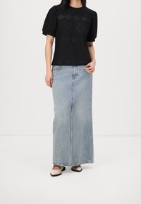 Blusa nera con texture e maniche a puff corte, abbinata a una lunga gonna di jeans azzurra. Modella che indossa scarpe beige con dettagli neri.